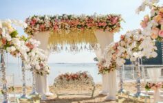 Elegant beach wedding setup in Bali featuring a white floral mandap with pastel roses, orchids, crystal chandeliers, and ocean view backdrop.