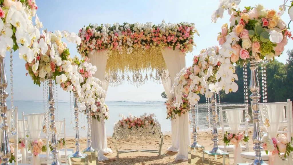 An extravagant luxury beach wedding altar decorated with white orchids, pink roses, and hanging crystal chandeliers overlooking the ocean, showcasing scenic places to visit in Bali.