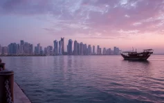 wide-angle landscape shot taken during sunrise of the high-rise skyscraper skyline of Doha, Qatar, across the water from the Corniche. A traditional wooden Dhow boat sails past with pink and purple clouds in the sky. Places to Visit in Doha