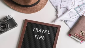 A flat lay featuring a vintage camera, a straw hat, a passport, a map, a toy plane, and a letterboard that reads travel tips.