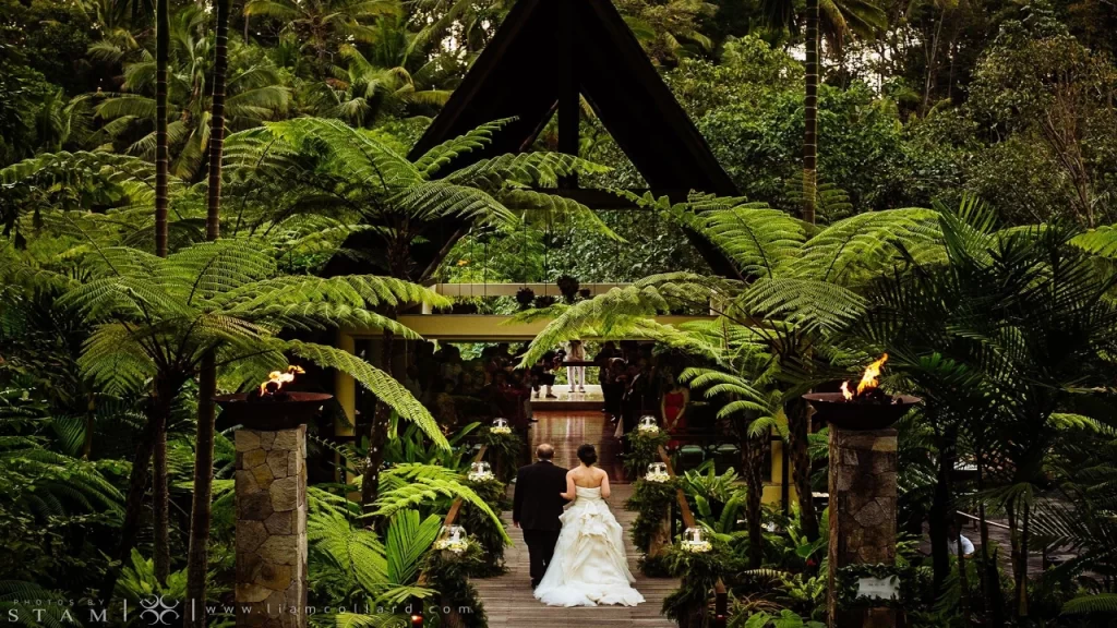 A bride and groom walking down a wooden aisle toward a striking A-frame jungle wedding chapel surrounded by lush tropical ferns and torches, highlighting unique places to visit in Bali.