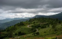 A winding asphalt road curving through lush green hills and pine trees with misty mountain ranges under dramatic storm clouds in the Knuckles region.