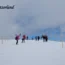 Must-Visit Tourist Places in Switzerland posing and taking photos on a vast snow-covered alpine summit in Switzerland, with red triangular warning STOP signs and rope barriers marking a safety boundary along the snowy ridge.