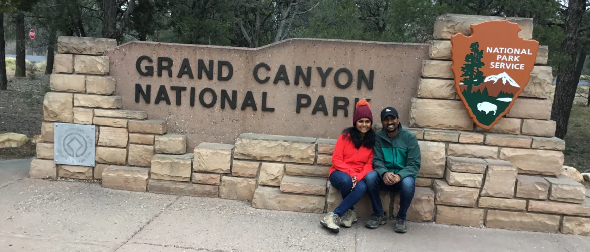 Grand Canyon National Park entrance sign with two visitors sitting beside the National Park Service sign surrounded by trees.