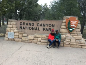 Grand Canyon National Park entrance sign with two visitors sitting beside the National Park Service sign surrounded by trees.