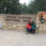 Grand Canyon National Park entrance sign with two visitors sitting beside the National Park Service sign surrounded by trees.